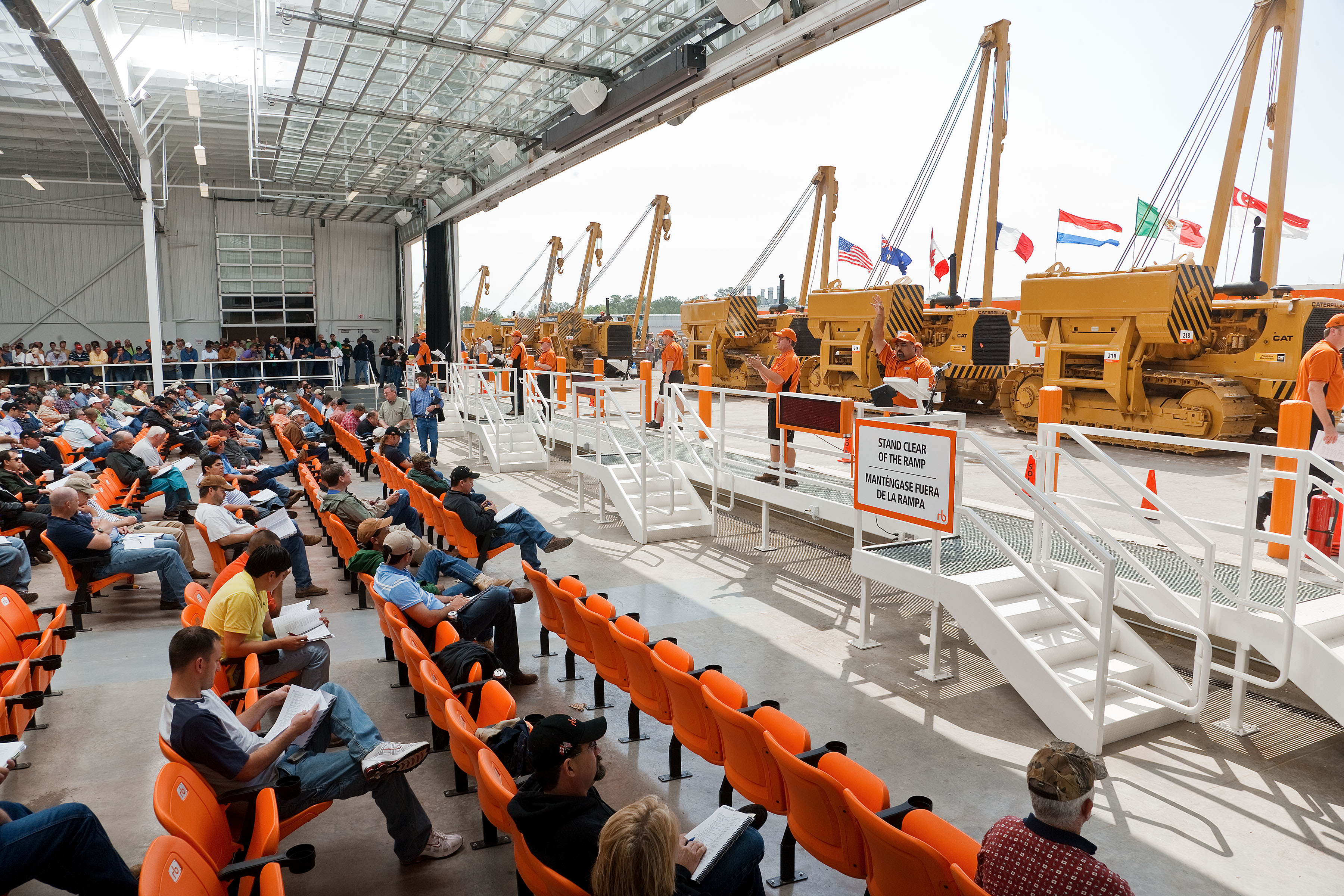 Auction site with a collection of heavy equipment on display and rows of bidders sitting in the audience.