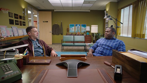 Still from Detroiters: Sam and Tim stare at each other puzzled on the same side of an executive desk.
