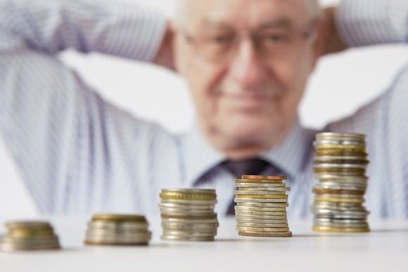 A man with his hands clasped behind his head looks smiling at progressively taller stacks of coins.