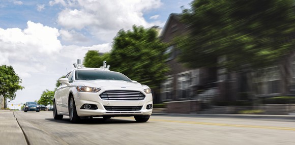 A white Ford Fusion sedan with autonomous-driving sensors visible, on a suburban road