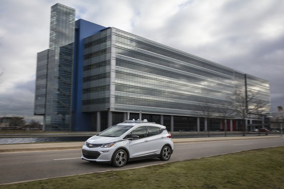 A white Chevrolet Bolt EV with self-driving sensors is shown on a public road near GM's technical center.