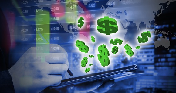 Green dollar signs floating in front of a screen with stock prices on it