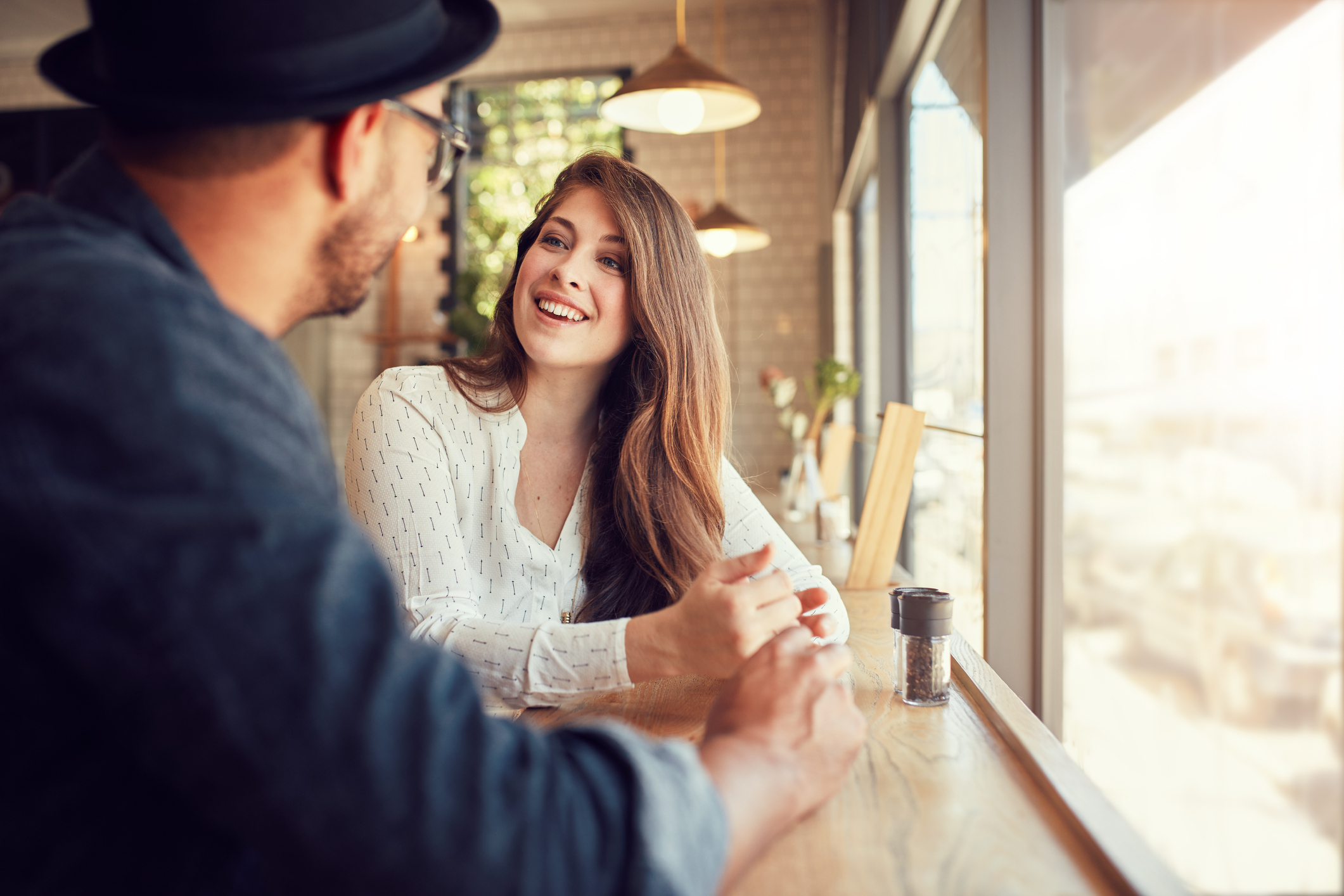 A smiling young woman sits in a cafe, talking to a man.
