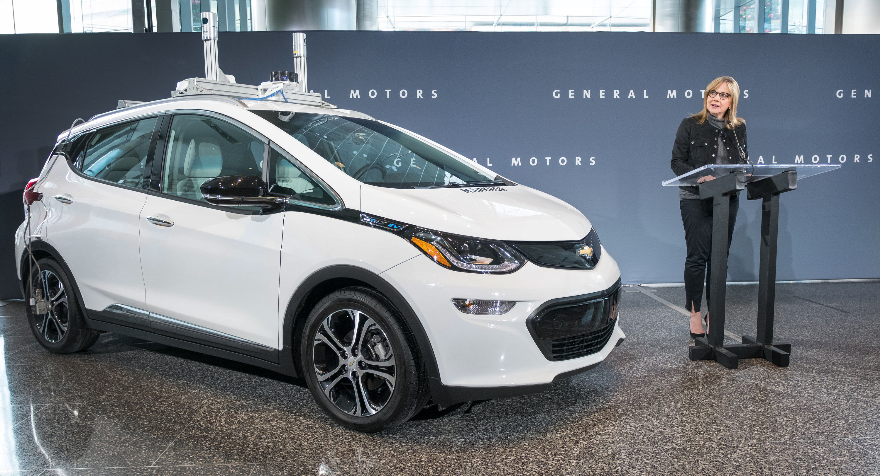 Barra is shown speaking while standing next to a white Chevrolet Bolt EV equipped with self-driving sensors.