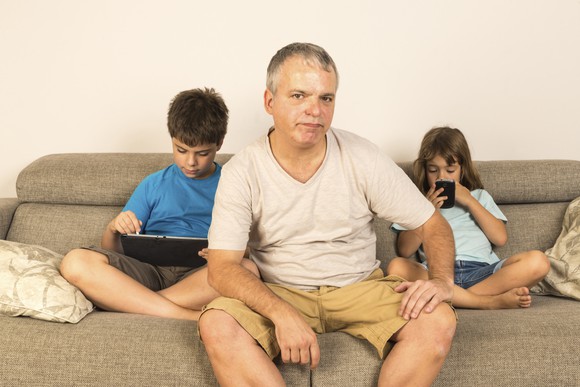 A man on a couch with two children, one looking at a tablet and another looking at a smartphone