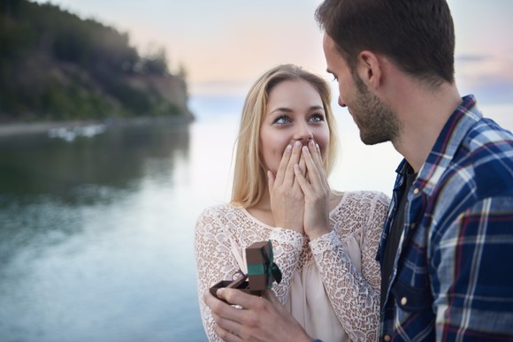 Young man proposing to surprised-looking girlfriend with ring in his hand