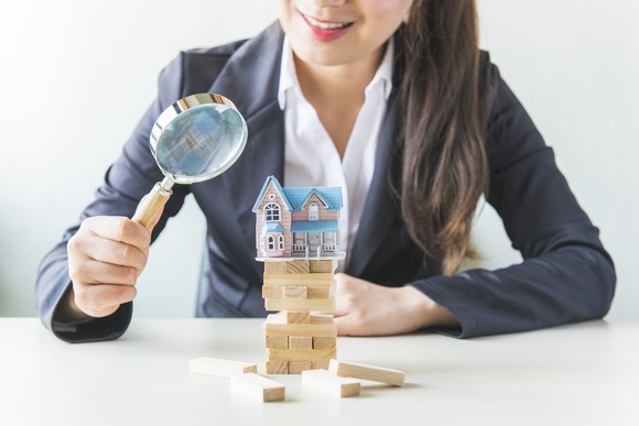 A woman looks through a magnifying glass at a model house piled on a stack of blocks.