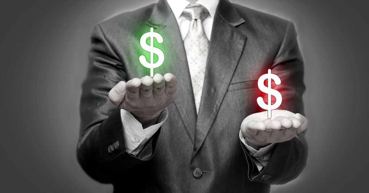 A business man holds a green dollar sign in one hand, slightly elevated above a red dollar sign held in his other hand.