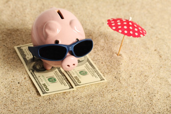 A piggy bank with sunglasses sits on two hundred-dollar bills, with a small umbrella, meant to resemble a beach umbrella, sitting next to it in the sand.