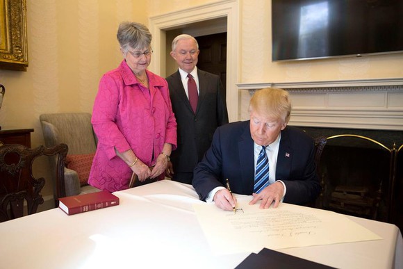President Trump signing paperwork while flanked by Attorney General Jeff Sessions and his wife.