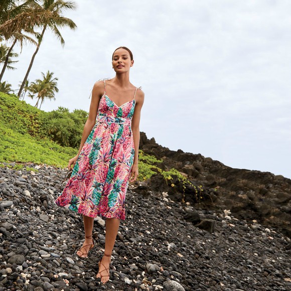 A woman stands on the rocks in a colorful dress, with the sky and a patch of grass behind her.