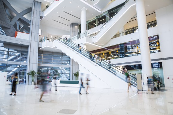 Shoppers walk through the inside of a mall, with an escalator in the background.