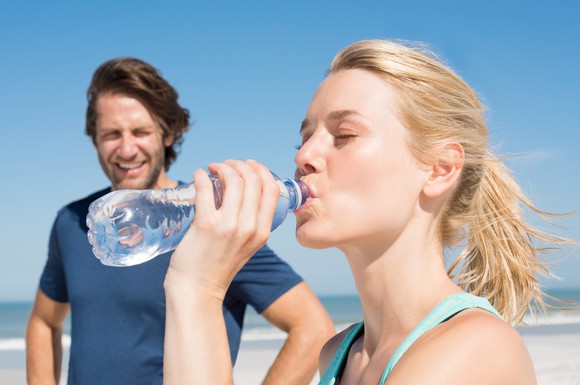 Woman runner drinking from water bottle on beach with male companion watching.