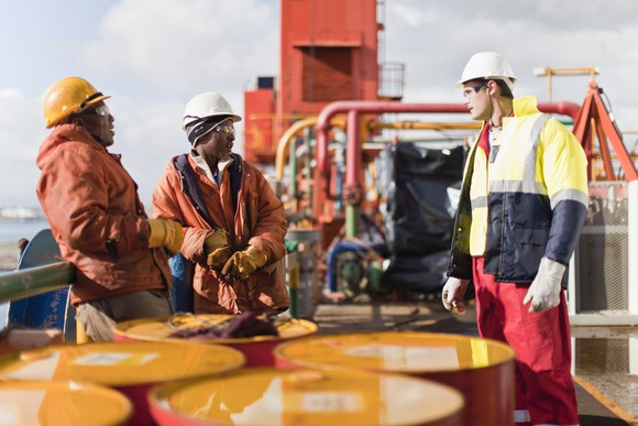 Three workers talk on an oil rig, with a group of oil barrels in the foreground.