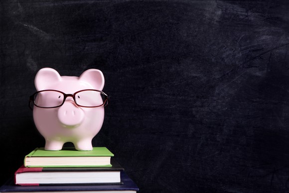 Wearing eyeglasses, a piggy bank stands on books in front of a chalkboard.