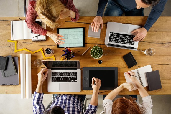 Team members sitting around a table with their laptops and working together.