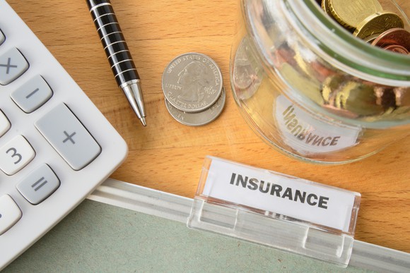 A folder labeled "insurance" sits on a desk, along with coins, a pen, and a calculator.