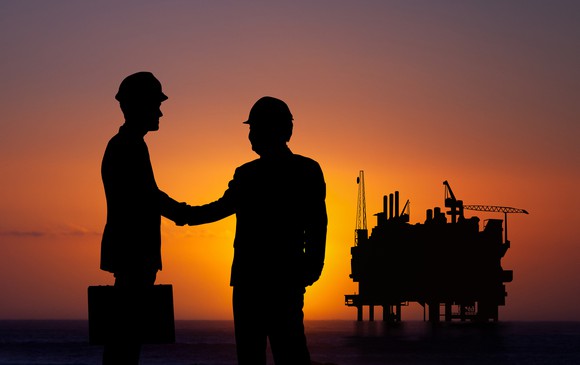 Two people in hard hats shaking hands near and oil and gas platform