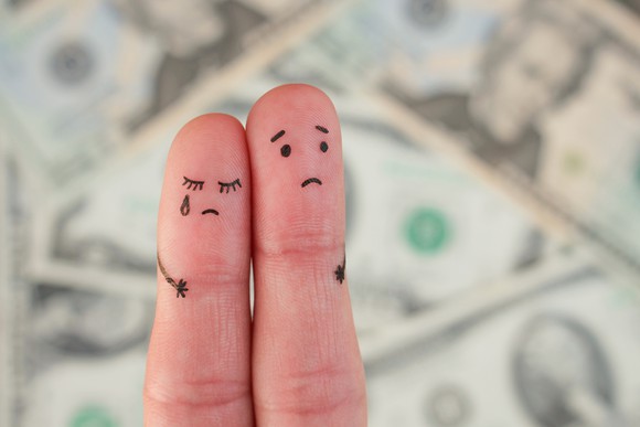 two fingers with sad faces drawn on them, in front of blurry dollar bills