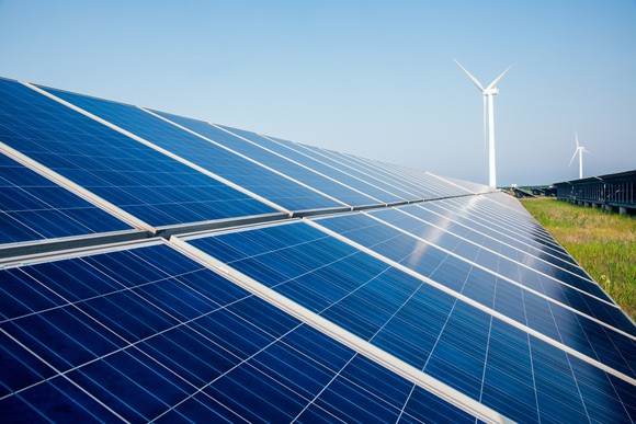 Array of solar panels with two wind turbines in the background on a sunny day.