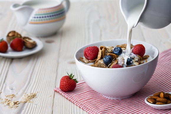 Milk is poured into a bowl of cereal on a tabletop, with other breakfast items nearby.
