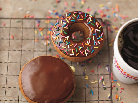 An overhead view of a cup of coffee and two doughnuts, both frosted with chocolate and one covered in colorful sprinkles.