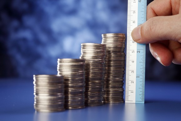 A hand holds a ruler, measuring stacks of coins growing progressively taller.