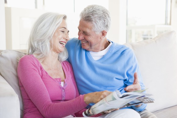 A retired couple sits smiling side by side on a sofa.