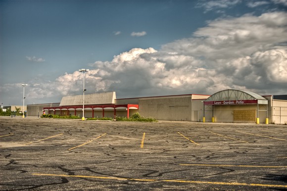 An abandoned shopping mall, as seen from across an empty, decaying parking lot.