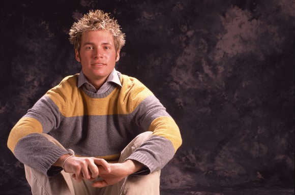 A crouched young man in a pullover displays preppy fashion style.