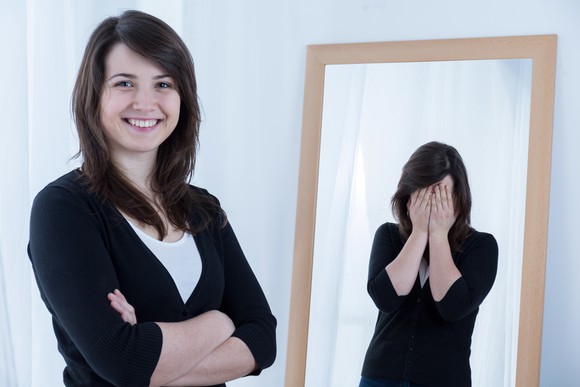 A woman looks confident, but in the mirror, she has her hands over her face.