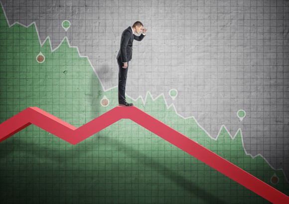 Business man looking downward while standing on a downward pointing arrow with a graph in background.