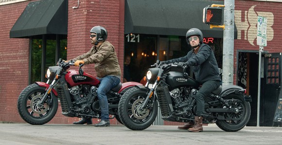 Two new Indian Scout Bobbers, one red, one black, with a male rider astride each, stopped outside a brick-building corner bar