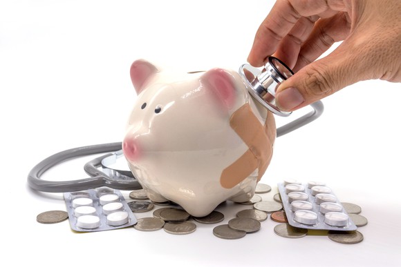 A person holds a stethoscope up to a piggy bank wearing band-aids.