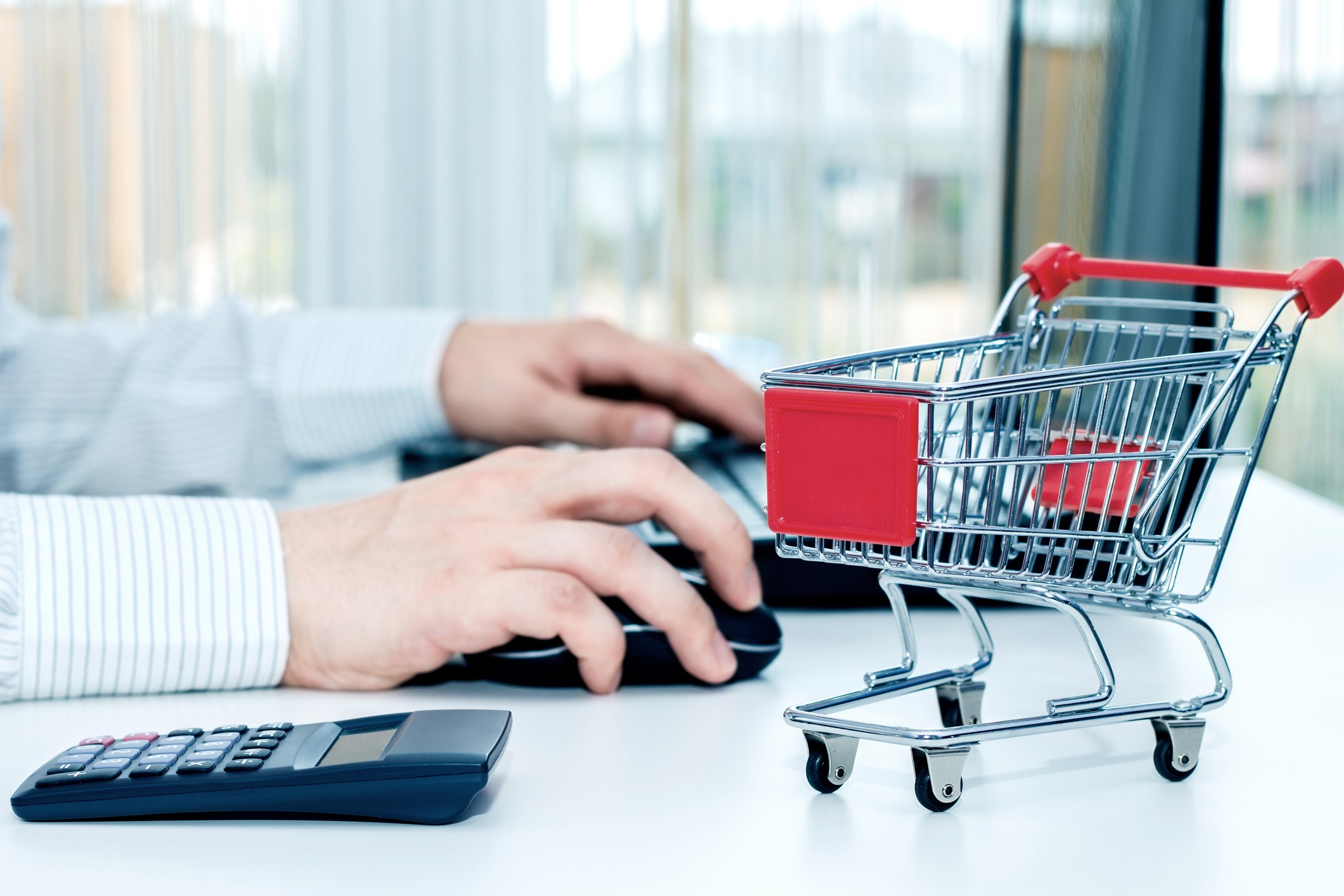 Man working at computer with calculator and a model of a shopping cart next to him.