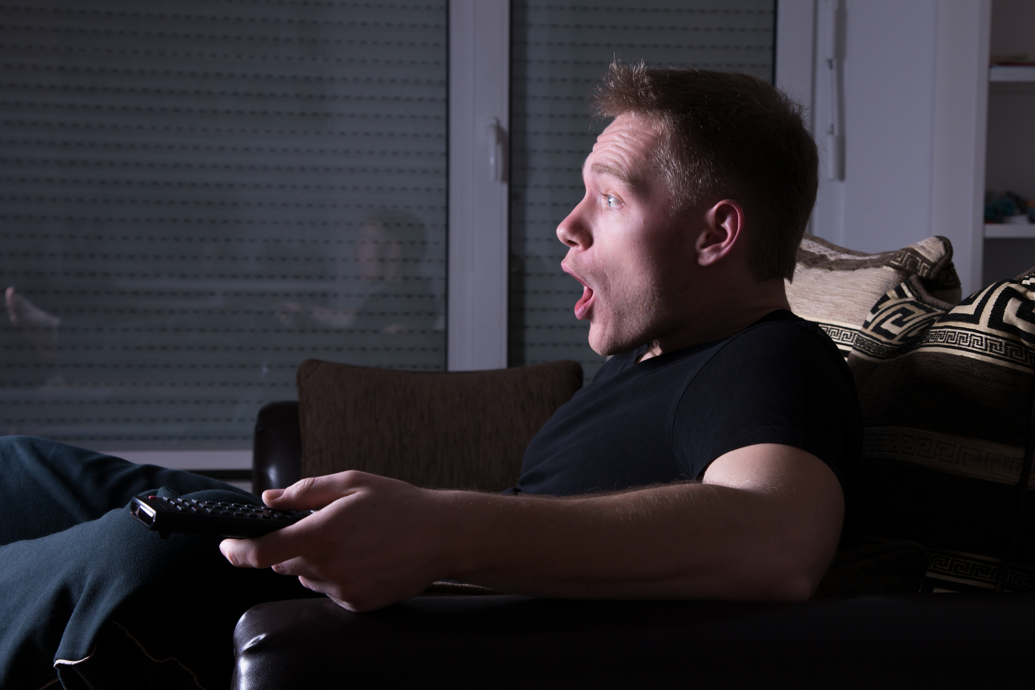 Man sitting in couch, visibly shocked by what's on the TV screen.