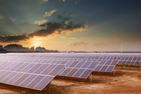 Solar farm at dusk with wind turbines in the background.