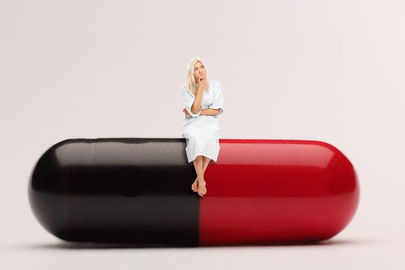 A woman contemplates a decision while sitting on a giant two-tone medicine capsule.