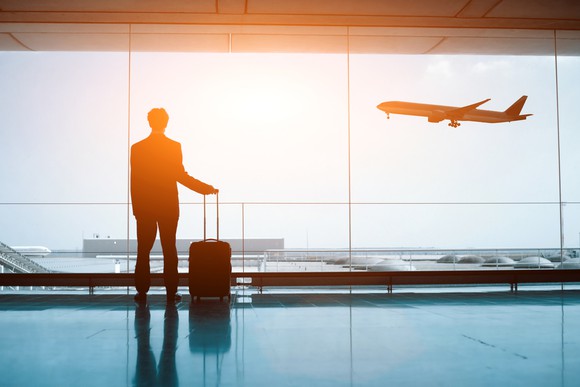 Silhouetted against the sun, a man stands with his luggage in an airport, watching through a window as an airplane takes off.