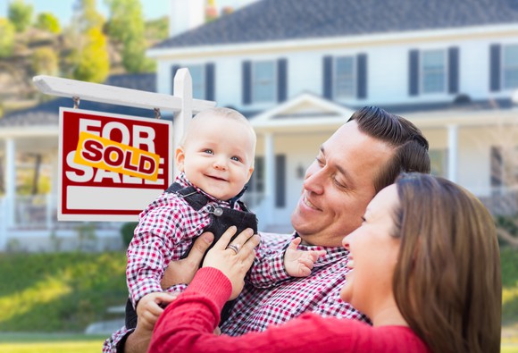A man and woman smile at their child in arms, next to a "sold" sign in front of a house.