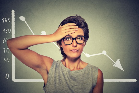 A woman slaps her forehead in front of a chart displaying a dropping stock price.