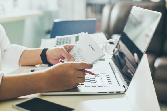 A person holds a resume and a pencil over a laptop.