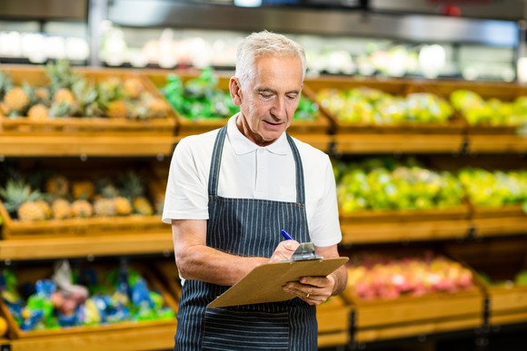 In a grocery-store setting, a gray-haired man in a short-sleeved shirt and apron writes on a clipboard.