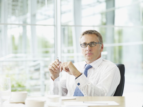 A business man wearing glasses sits behind his desk in front of a glass wall.