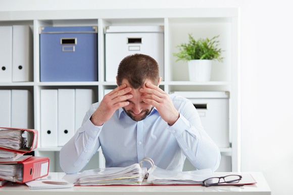 A man looks down at papers in an open binder, with his fingers pressed into his forehead, suggesting frustration.