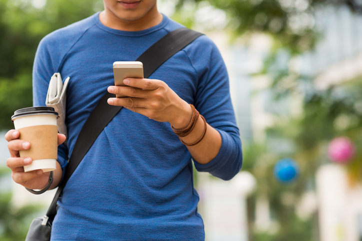 Young man walking with mobile phone, coffee, and newspaper tucked under arm.