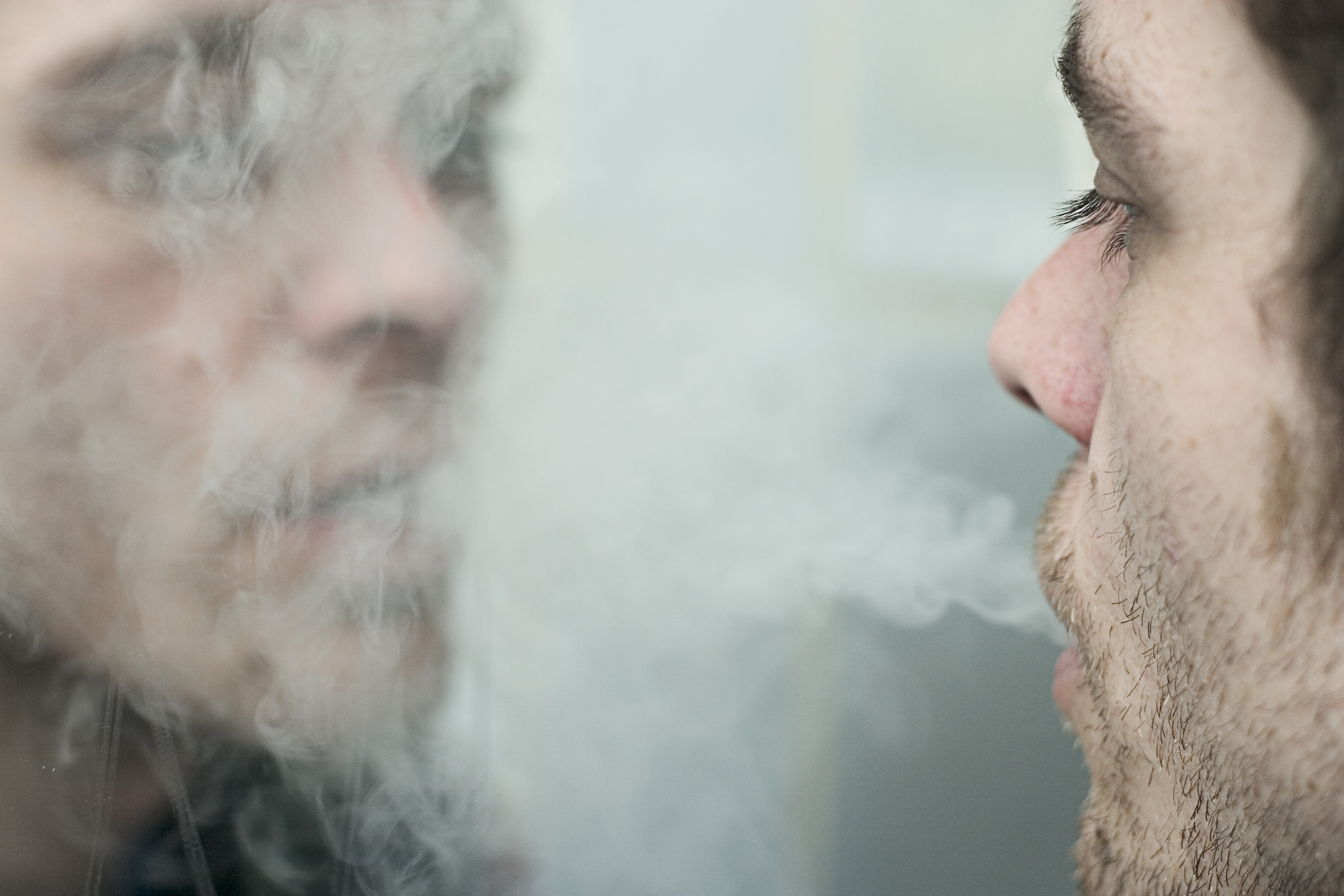 A man blows cigarette smoke at his reflection in the mirror.