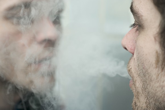A man blows cigarette smoke at his reflection in the mirror.