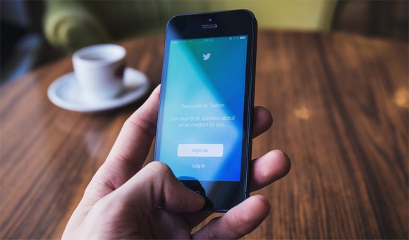 A hand holds a smartphone over a wooden table, with a cup and saucer in the background. On th screen is the Twitter app.