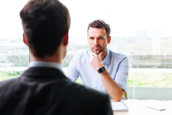 A man, hand to his chin, looks across a table at an interviewee.